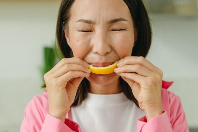 Asian woman in pink shirt makes a funny face while tasting a sour lemon slice indoors. Une femme asiatique en chemise rose fait une grimace amusante en goûtant une tranche de citron acide à l'intérieur.