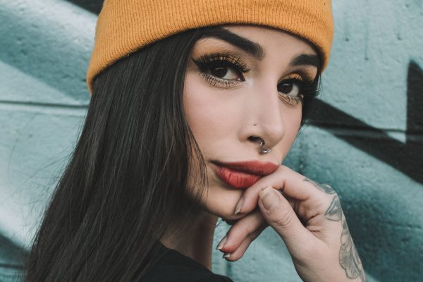 Close-up portrait of a woman in a yellow beanie, displaying tattoos and nose piercing. Portrait en gros plan d'une femme portant un bonnet jaune, affichant des tatouages et un piercing au nez.