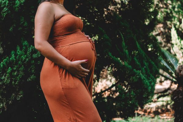 A pregnant woman standing and smiling in a lush outdoor setting, captured in a warm portrait. Une femme enceinte debout et souriante dans un cadre extérieur luxuriant, capturée dans un portrait chaleureux.